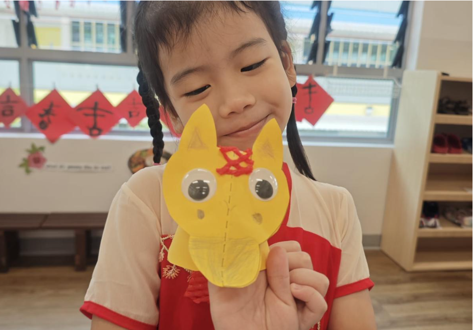 Girl in a red dress holds a yellow paper cat craft with googly eyes in a classroom.